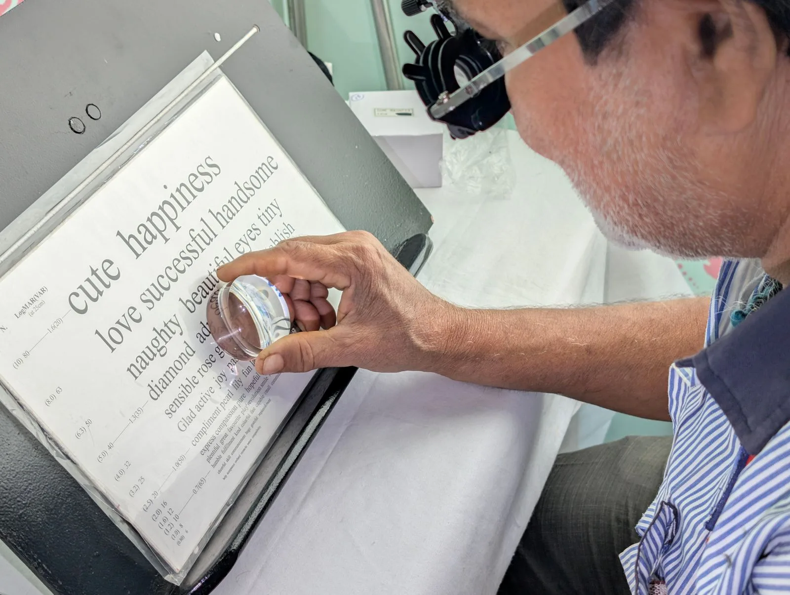 An older man in a trial frame reading a near-vision chart with a magnifier during an eye test.