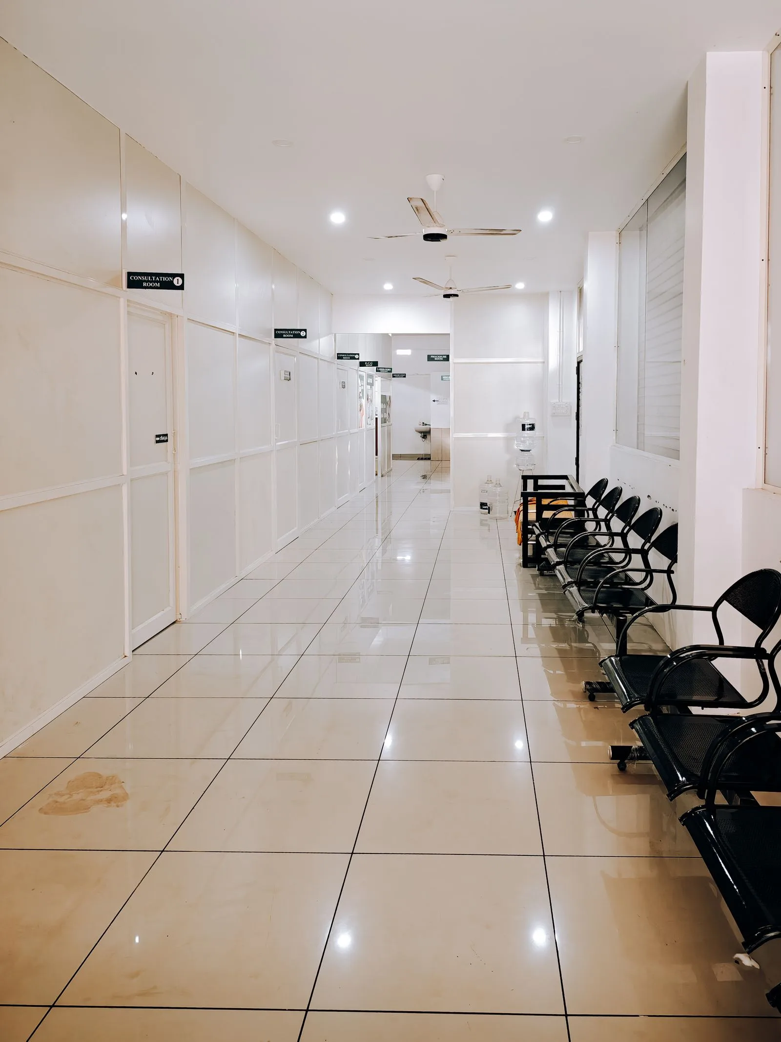 A bright hospital corridor with a row of waiting chairs outside the consultation rooms.